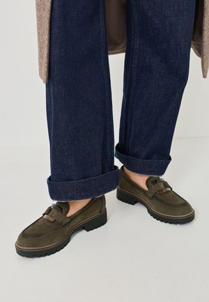 Henkilö, joka käyttää tummansinisiä käännettyjä farkkuja ja oliivinvihreitä mokkanahkaisia loafer-kenkiä paksuilla mustilla pohjilla, seisoo vaalealla lattialla.
