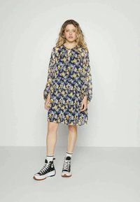 Jovem mulher vestindo um vestido floral azul marinho com mangas longas transparentes e tênis de cano alto preto e branco, em pé sobre um piso cinza.