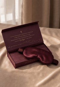 Seidene, dunkelviolette Augenmaske, die auf einer geöffneten passenden Schachtel mit goldener Schrift auf einem beigen Stoffhintergrund liegt.