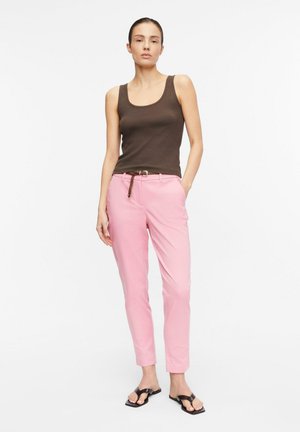 Kobieta stojąca, ubrana w bezrękawnik ciemnobrązowy top, jasnoróżowe spodnie z paskiem i czarne sandały z odkrytymi palcami na jednolitym białym tle.