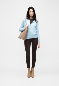 Γυναίκα που στέκεται φορώντας ένα ανοιχτό μπλε πλεκτό ζακέτα με κουμπιά, σκούρο καφέ παντελόνι, ταμπά μποτάκια μέχρι τον αστράγαλο και κρατώντας μια μεγάλη μπεζ τσάντα ώμου.