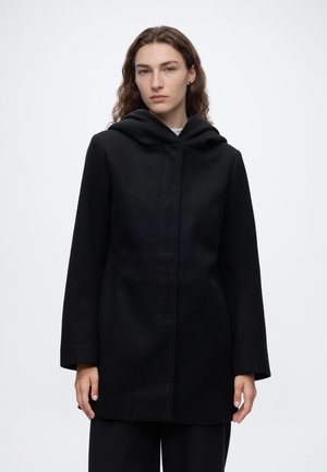 Personne aux cheveux longs portant un manteau à capuche noir et un pantalon noir, debout devant un fond clair uni avec une expression neutre.