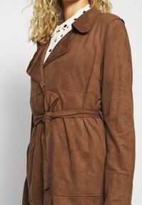 Manteau en daim marron avec une taille ceinturée, un large col et une fermeture à boutons. Il présente une texture lisse et des poches latérales pour plus de détails.