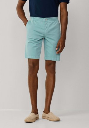 Mann trägt hellblaue Shorts mit elastischem Bund, Knopf und Reißverschluss, ein marineblaues Hemd und beige Slip-on-Espadrilles, steht vor einem einfarbigen Hintergrund.