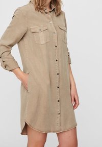 Beige skjortklänning i mjukt tyg, med två bröstfickor, uppvikta ärmar och rundad nederkant.