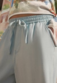 Helle blaue, elastische Hose mit einem Kordelzug, die eine glatte Textur und Seitentaschen aufweist. Kombiniert mit einem bunten gemusterten Hemd.