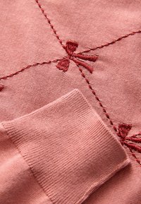 Rosa gestrickter Stoff mit roten bestickten Schleifen und Nähten. Verfügt über einen gerippten Bund und eine glatte Textur, die aufwändige Details zeigt.