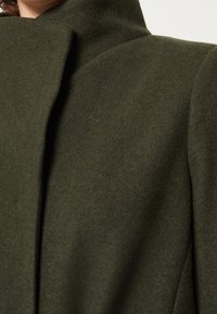 Olive green wool-blend coat with a high, structured collar and a smooth, matte texture. Features tailored seams and a fitted silhouette.