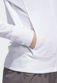 Jaqueta branca com fecho à frente, feita de tecido texturizado com um padrão geométrico, mangas longas e um bolso frontal. Design suave e leve.