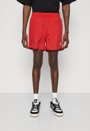 Red athletic shorts with an elastic waistband and white side stripes. Worn with black and white sneakers and white socks.