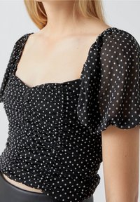 Schwarzes Oberteil mit weißen Punkten, gerafftem Stoff, quadratischem Ausschnitt und transparenten Puffärmeln. Strukturierter Stoff mit weißen Punkten sorgt für einen spielerischen Kontrast.