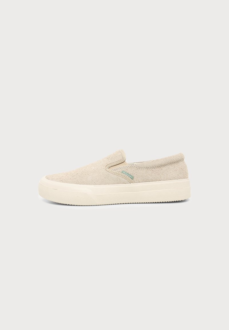 Ανοιχτό μπεζ αθλητικό παπούτσι slip-on με υφασμάτινο επάνω μέρος με υφή και χοντρή λευκή καουτσούκ σόλα σε μονόχρωμο φόντο.