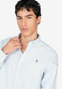 Lichtblauw geblokt button-up shirt met een kraag, voorzien van witte knopen en een klein marineblauw logo op de borst. Smooth stoftextuur.