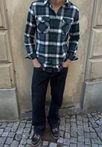 Person som bär en grön och blå rutig skjorta med knappar, mörka jeans och bruna läderstövlar står på kullersten nära en beige vägg.