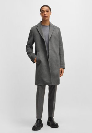 Grau Wollmantel mit Reverskragen, zweiknöpfigem Vorderteil und seitlichen Taschen. Kombiniert mit grauen Hosen und schwarzen klobigen Stiefeln.