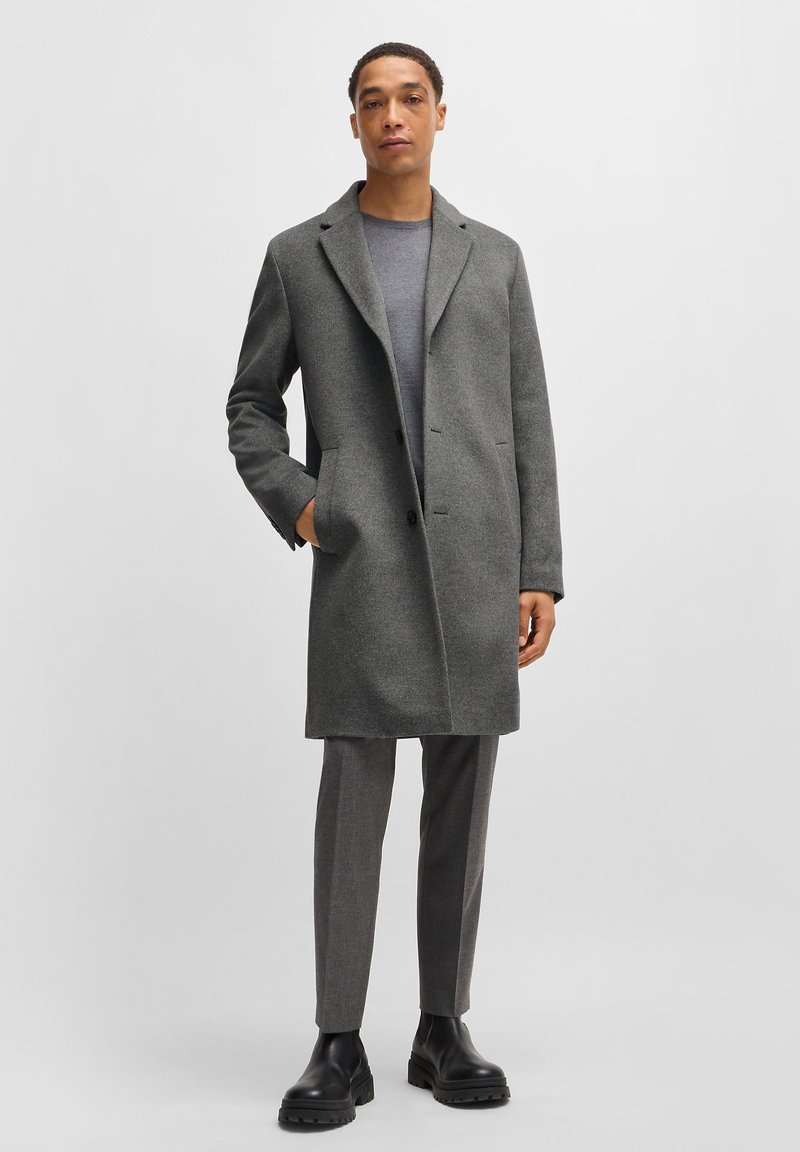 Grau Wollmantel mit Reverskragen, zweiknöpfigem Vorderteil und seitlichen Taschen. Kombiniert mit grauen Hosen und schwarzen klobigen Stiefeln.