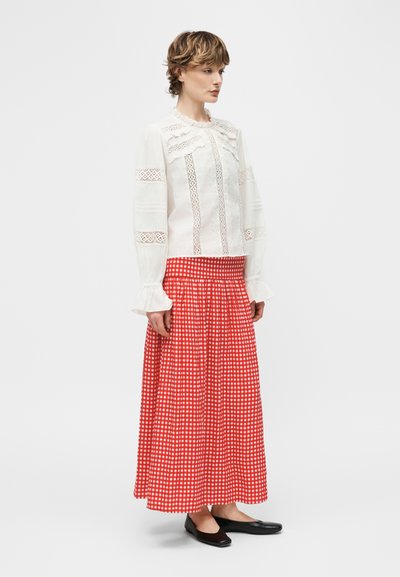 Άτομο που στέκεται φορώντας μια λευκή μπλούζα με λεπτομέρειες από δαντέλα, μακριά φούστα με κόκκινο και λευκό καρό σχέδιο και μαύρες μπαλαρίνες σε απλό λευκό φόντο.