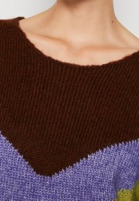 Close-up of a person wearing a knitted sweater with brown, purple, and green color blocks and a wide neckline.