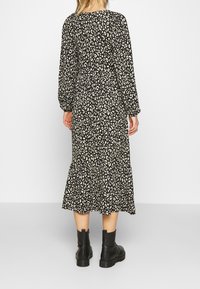 Femme portant une robe midi à manches longues à imprimé léopard noir et blanc avec des bottines noires, debout devant un fond blanc uni.