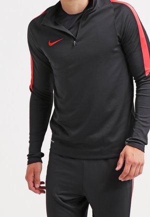 Homme portant un haut de sport noir à manches longues avec des rayures rouges sur les manches et un logo Nike rouge sur la poitrine.