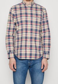 Man draagt een geruit overhemd met lange mouwen en knopen, met rode, blauwe en gele strepen, gecombineerd met een blauwe spijkerbroek, staat met ontspannen armen.