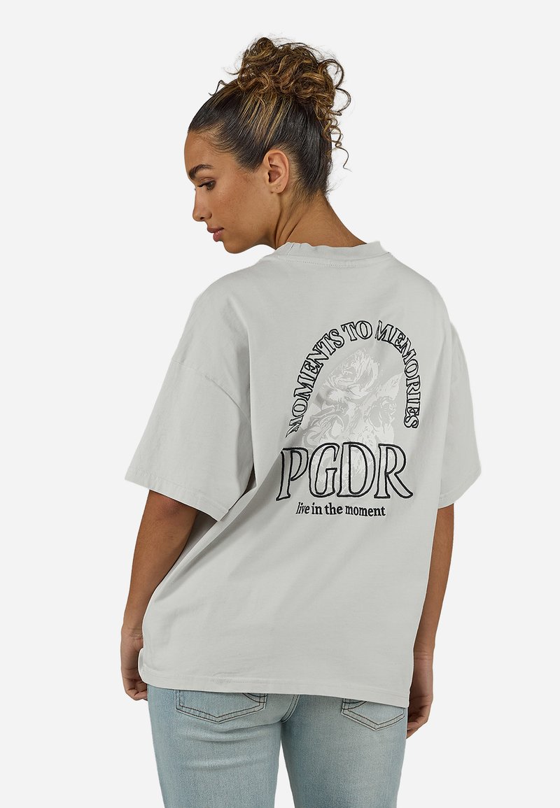 Γυναίκα με σγουρά μαλλιά δεμένα ψηλά φοράει μια φαρδιά ανοιχτό γκρι μπλούζα με το κείμενο «Moments to Memories PGDR live in the moment» και floral σχέδιο στην πλάτη.