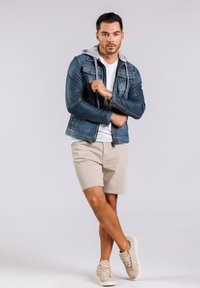 Blaue Lederjacke mit grauer Kapuze, weißem T-Shirt, beigen Shorts und beigen Sneakern. Das Model posiert mit verschränkten Armen vor einem neutralen Hintergrund.