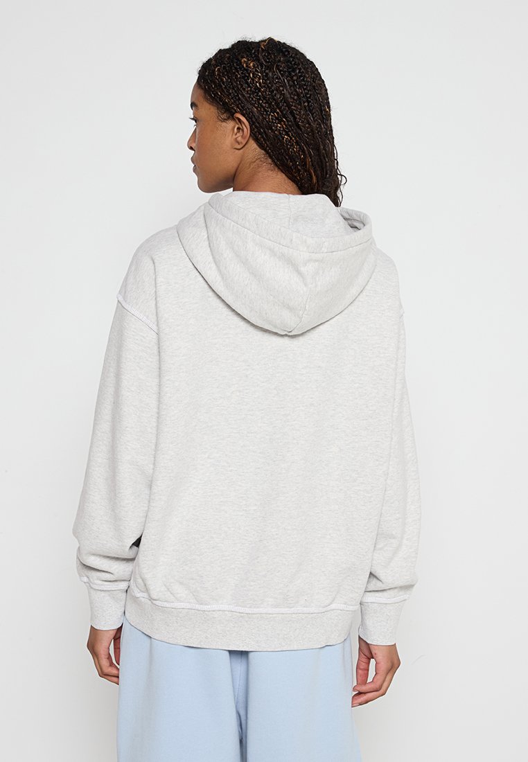 Frau mit geflochtenem Haar, die einen hellgrauen Hoodie und hellblaue Hosen trägt, von hinten vor einem schlichten weißen Hintergrund gesehen.