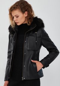 Giacca in pelle nera con cappuccio bordato in pelliccia, chiusure con zip e bottoni a pressione, due tasche sul petto e una finitura liscia. Design semplice e su misura.