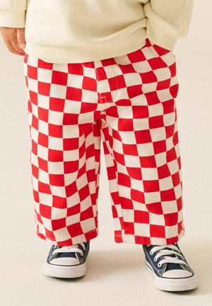 Kind trägt rot-weiß karierte Hose, marineblaue Sneakers mit weißen Schnürsenkeln und einen cremefarbenen Pullover.