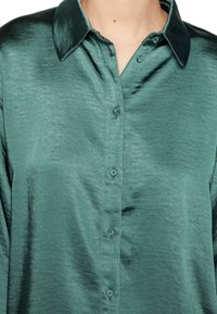 Camisa de botones en satén verde oscuro, con un cuello clásico, ligero brillo y seis botones en la parte delantera, con una textura suave.