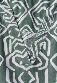 Tela verde con patrones geométricos blancos, con escote en V y detalle de costura visible en los bordes. Textura suave y lisa.