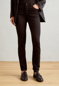 Zwarte slim-fit broek met een gladde textuur en vijf zakken, gecombineerd met zwarte instappers op een houten vloerachtergrond.