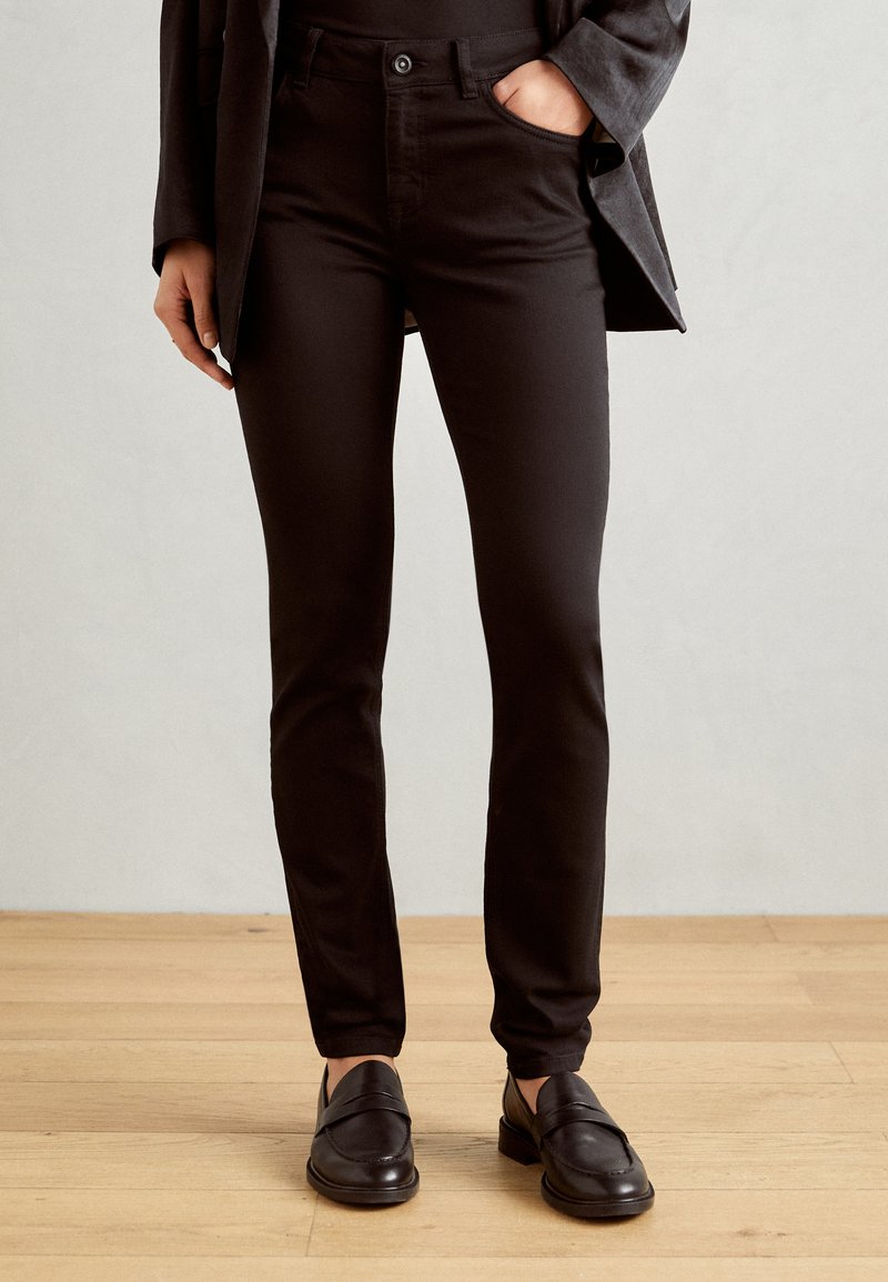 Zwarte slim-fit broek met een gladde textuur en vijf zakken, gecombineerd met zwarte instappers op een houten vloerachtergrond.