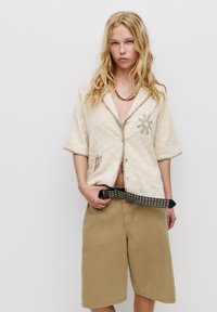 Cămașă bej cu mâneci scurte, decorată cu broderie florală și închidere cu nasturi, asortată cu pantaloni scurți bej și un curea neagră cu tinte. Fundal neutru.