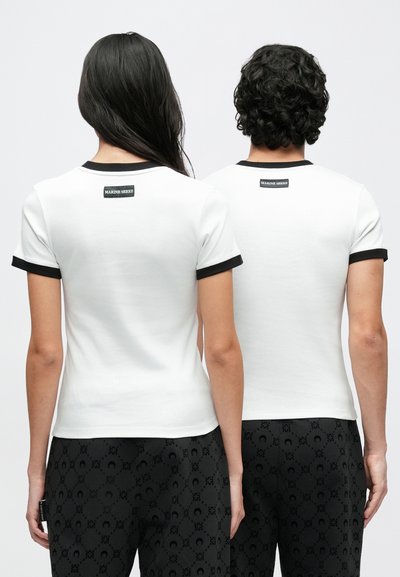 Κομμάτια λευκών βαμβακερών μπλουζών με κοντά μανίκια, μαύρα ριμπ κολάρα και ετικέτα στο πίσω μέρος. Συνδυασμένα με μαύρο παντελόνι με σχέδια.