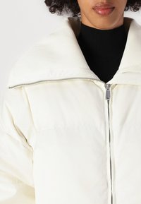 Dunhvitt jakke med ribbet kremfarge krage og sølvglidelås. Båret over en svart turtleneck, som viser en glatt, blank overflate.