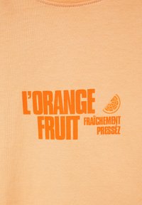 Texte orange indiquant "L'ORANGE FRUIT FRAÎCHEMENT PRESSÉ" avec une petite illustration d'une tranche d'orange sur un tissu orange clair.