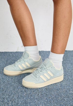 Nogi w białych skarpetach typu crew i jasnoniebieskich butach sportowych Adidas z kremowymi paskami i podeszwami na niebieskim dywanie.