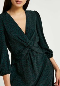 Vestido de estampado de leopardo verde con un escote en V profundo, detalle en el delantero retorcido y mangas abullonadas. Hecho de una tela suave y texturizada.