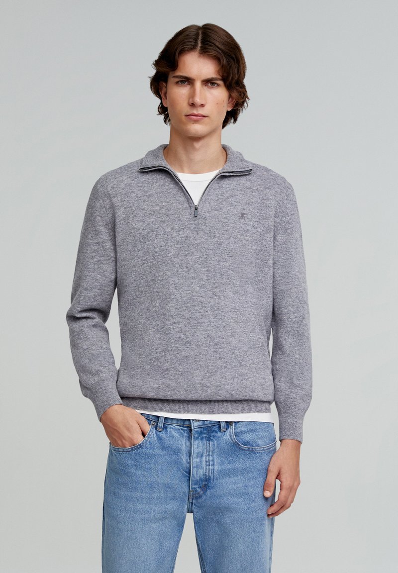 Grauer Strickpullover mit einem Reißverschluss und Stehkragen, gerippten Bündchen und Saum, getragen über einem weißen T-Shirt und kombiniert mit hellblauen Jeans.