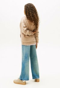 Persona con cabello largo y rizado de espaldas, vistiendo una sudadera beige, jeans anchos de mezclilla azul desgastados y zapatillas beige, sobre un fondo blanco.