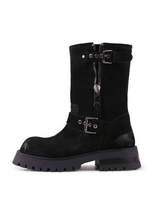 Botas de tobillo de ante negro con suela resistente, que cuentan con un cierre lateral, correa con hebilla y áreas de acentos brillantes. Tacón plano para mayor estabilidad.