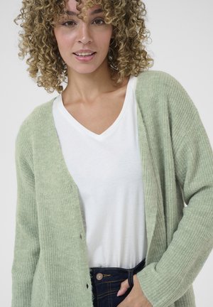 Light green knitted cardigan with a loose fit and open front, worn over a white V-neck t-shirt. Features ribbed texture and button accents.