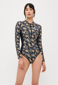 Blomstrete badedrakt med lange ermer som har svart bakgrunn med gule, blå og oransje blomster; glidelås foran, atletisk snitt og glatt tekstur.
