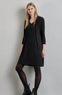 Robe plissée noire avec un décolleté en V et des manches trois-quarts, associée à des bottines noires et des collants noirs semi-transparents sur un fond uni.