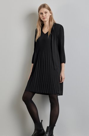 Robe plissée noire avec un décolleté en V et des manches trois-quarts, associée à des bottines noires et des collants noirs semi-transparents sur un fond uni.