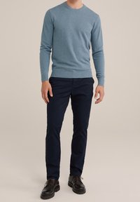 Man i ljusblå stickad tröja, mörka marinblå byxor och svarta läder snörskor, stående mot en neutral bakgrund.