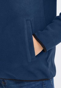 Giacca in pile navy con tasca laterale con zip, rifiniture nere sui polsini e una texture morbida. Dettaglio del lato sinistro.