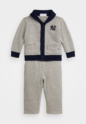 Pelēks adīts divdaļīgs tērps, kas sastāv no pogājamas kardigānas ar tumši zilām šallēm un logo, apvienots ar atbilstošām biksēm. Mīksta faktūra visā apģērbā.